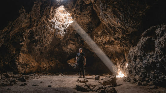 A person stands in a large rocky cave, illuminated by a beam of sunlight streaming down through an opening in the ceiling. The cave interior is rugged with scattered rocks on the ground. by Justin Salem Meyer