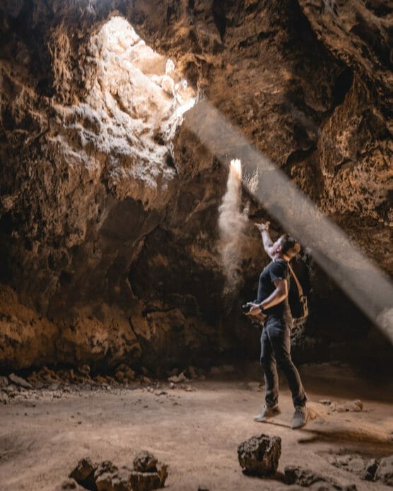A man standing inside a cave tosses dust into a sunbeam coming through an opening above, illuminating the dust in the air. He is holding a camera and looking up toward the light. Rocky cave walls surround him. by Justin Salem Meyer