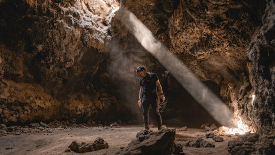 A person standing in a rocky cave is illuminated by a beam of sunlight streaming in from an opening above, creating a dramatic contrast between light and shadow on the cave walls and floor. by Justin Salem Meyer