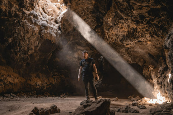A person stands in a rocky cave illuminated by a beam of sunlight streaming through an opening above, highlighting dust in the air and casting dramatic shadows on the cave walls. by Justin Salem Meyer