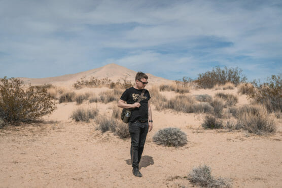 A man in a black t-shirt and sunglasses walks through a sandy desert landscape with sparse shrubs and distant hills under a partly cloudy sky. by Justin Salem Meyer