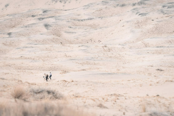 Three people walk across a vast, sandy desert landscape with rolling dunes and sparse vegetation, highlighting the scale and emptiness of the environment. by Justin Salem Meyer