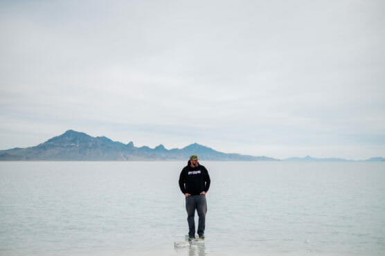 A person wearing a black hoodie and cap stands alone in shallow water with distant mountains and a cloudy sky in the background. by Justin Salem Meyer