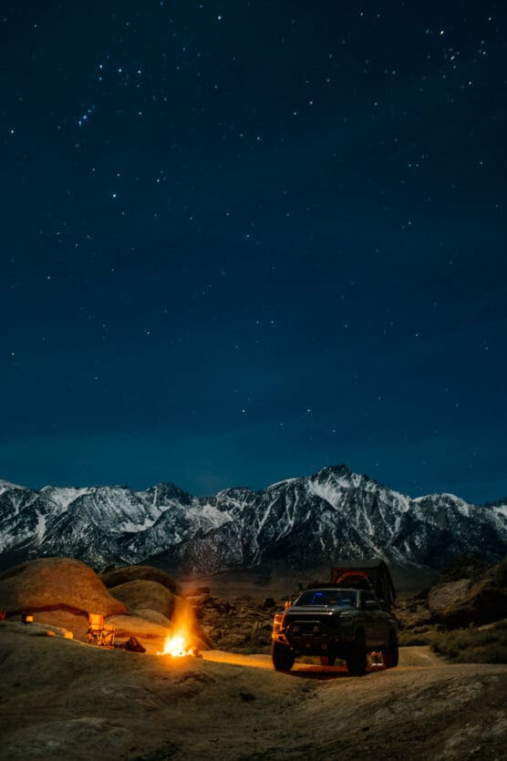 A campsite with a campfire, two chairs, and a vehicle under a starry night sky, set against snow-capped mountains and large rocks. by Justin Salem Meyer