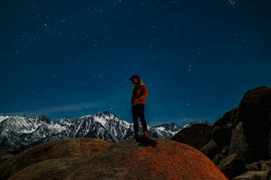 A person stands on a large rock at night, gazing at the star-filled sky, with snow-capped mountains and rugged rocks in the background. The scene is illuminated by soft, natural and warm light. by Justin Salem Meyer
