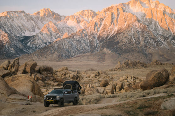 A gray off-road truck with a rooftop tent is parked among large boulders in a desert landscape, with tall, snow-capped mountains lit by the setting sun in the background. by Justin Salem Meyer