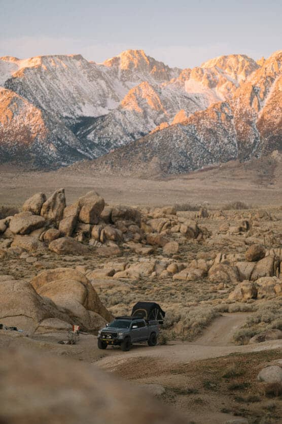 A black off-road vehicle with a rooftop tent is parked on a dirt path among large boulders and desert brush. Snow-capped mountains are illuminated by soft golden sunlight in the background. by Justin Salem Meyer