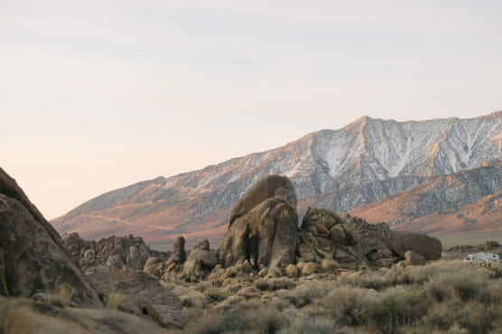 Large rocky boulders and dry grass in the foreground with snow-dusted mountains and a cloudy sky in the background, captured during soft, warm lighting. by Justin Salem Meyer