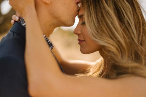 A close-up of a couple embracing outdoors, with one person gently kissing the others forehead. Both appear peaceful and affectionate, with soft sunlight illuminating their faces and hair. by Justin Salem Meyer