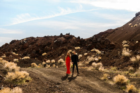 A woman in a bright red dress and a man in dark clothing walk side by side on a dirt path surrounded by rocky, desert-like terrain with sparse dry bushes under a partly cloudy sky. by Justin Salem Meyer
