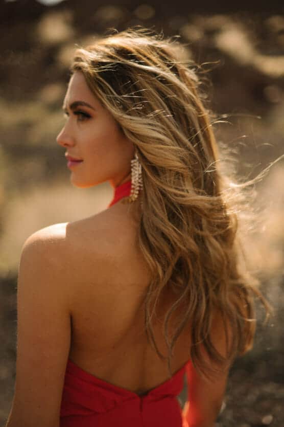 A woman with long, wavy blonde hair wears a red, backless dress and dangling earrings, standing outdoors in soft, natural light with a blurred, earthy background. by Justin Salem Meyer