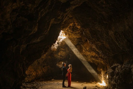 A couple stands facing each other in a large, rocky cave as a beam of sunlight streams down through an opening above, illuminating them and casting dramatic shadows on the cave walls. by Justin Salem Meyer