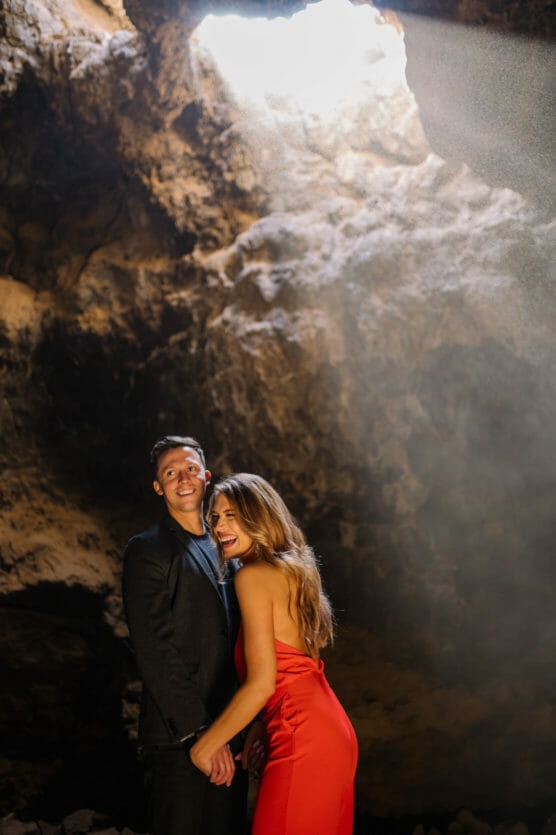 A smiling couple stands in a sunlit cave. The man wears a dark suit, and the woman wears a red dress. Sunlight streams through an opening above, illuminating them against the rocky background. by Justin Salem Meyer
