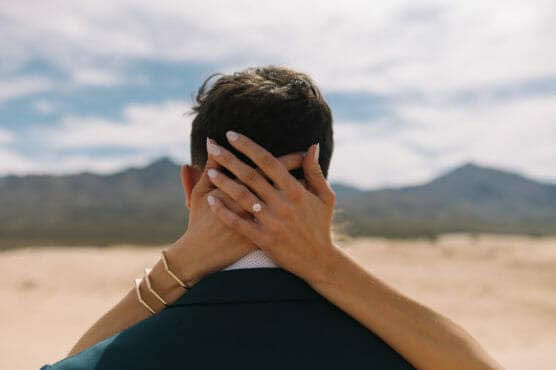 A person embraces a man from behind, hands resting on the back of his head and neck. The focus is on a diamond engagement ring and gold bracelets, with a desert landscape and mountains in the background. by Justin Salem Meyer