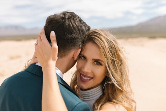 A woman smiles at the camera while hugging a man in a suit in a sunny, sandy desert landscape. She has long, blonde hair and wears a sleeveless turtleneck top. The man faces away from the camera. by Justin Salem Meyer