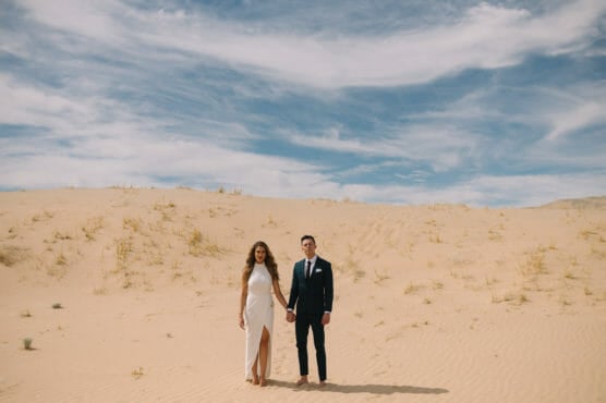 A couple dressed in formal attire—a woman in a white dress and a man in a dark suit—stand holding hands on a sandy desert landscape under a partly cloudy blue sky. by Justin Salem Meyer