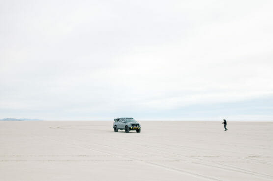 A person stands on a vast, flat, sandy landscape near a dark SUV under a cloudy sky. The area appears empty and expansive, with faint tire tracks visible on the ground. by Justin Salem Meyer