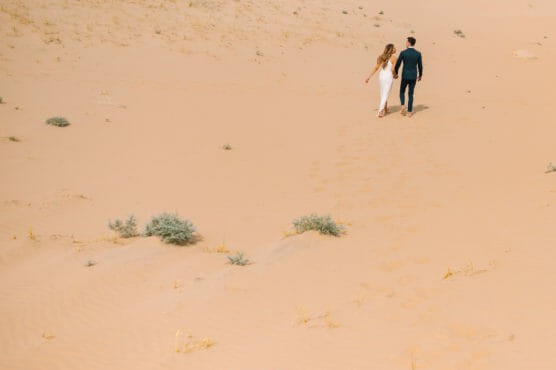 A couple walks barefoot together across a sandy desert landscape, leaving footprints behind them. Sparse green shrubs dot the sand, and the sky is not visible. by Justin Salem Meyer