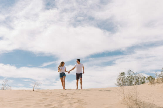 A couple holding hands walks on a sandy dune under a partly cloudy sky, with some small bushes in the background. by Justin Salem Meyer