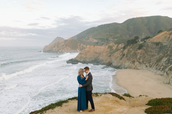 A couple embraces on a cliff overlooking the ocean, with waves crashing on a sandy beach below and green hills in the background under a partly cloudy sky. by Justin Salem Meyer