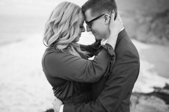 A couple embraces closely on a beach, touching foreheads and smiling softly. The image is black and white, and the background is blurred, highlighting their intimate connection. by Justin Salem Meyer
