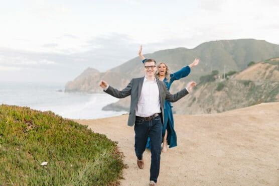 A man in a gray blazer walks on a coastal path with a woman in a blue dress behind him, both smiling and playfully holding their arms up. Green hills and the ocean are visible in the background. by Justin Salem Meyer