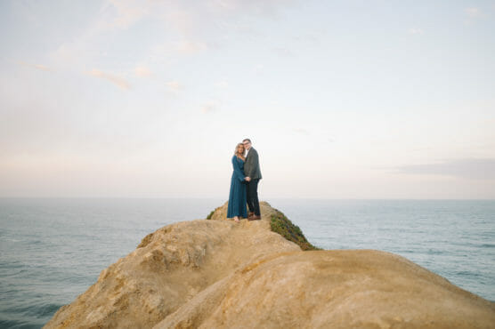 A couple stands together on a rocky cliff overlooking the ocean, with a soft, pastel sky in the background. They appear to be embracing, dressed in formal attire, surrounded by natural scenery. by Justin Salem Meyer