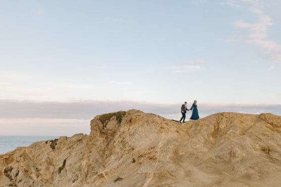 A couple holding hands walks along the top of a rocky cliff under a pale blue sky, with the ocean visible in the background. by Justin Salem Meyer