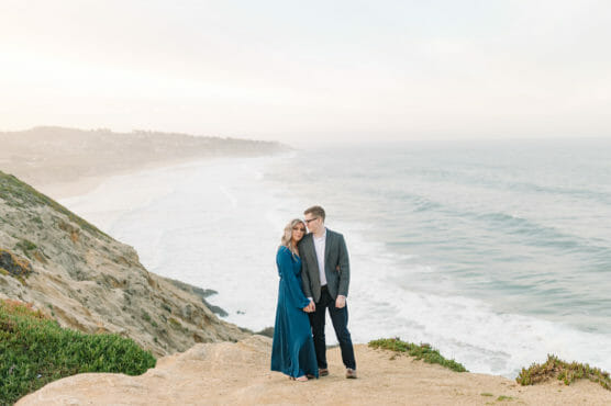A couple stands on a sandy cliff overlooking the ocean, with waves crashing below and distant hills in the background. The woman wears a long blue dress, and the man wears a jacket and jeans. The sky is bright and slightly hazy. by Justin Salem Meyer