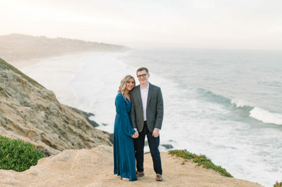 A couple stands on a rocky cliff overlooking the ocean, holding hands and smiling. The woman wears a long blue dress and the man wears a gray jacket and dark pants. Waves crash on the shore below under a cloudy sky. by Justin Salem Meyer