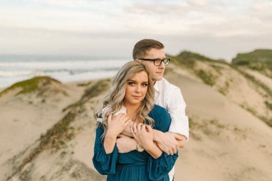 A couple stands on sandy dunes by the ocean. The man in glasses, wearing a white shirt, hugs the woman in a blue dress from behind. Both have a calm, serious expression. The dunes and cloudy sky are in the background. by Justin Salem Meyer