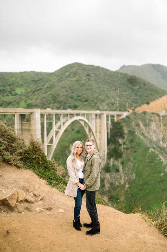 A couple stands smiling and holding hands on a dirt path with lush green hills and a large arched bridge in the background under a cloudy sky. by Justin Salem Meyer