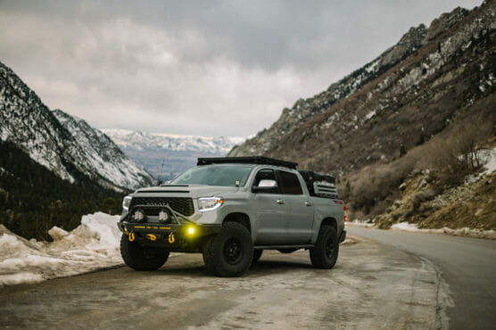 A silver pickup truck with off-road tires and roof rack is parked on a mountain road surrounded by snow and rocky hills under a cloudy sky. by Justin Salem Meyer