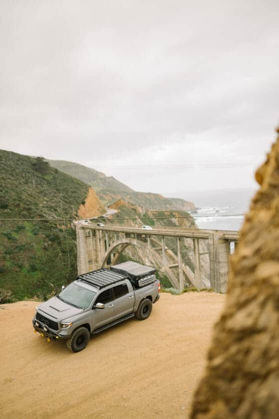 A gray off-road truck is parked on a dirt overlook near a scenic coastal bridge with green hills and the ocean in the background under a cloudy sky. by Justin Salem Meyer