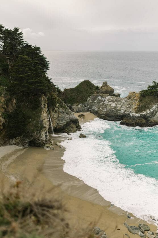 A secluded sandy beach with turquoise waves, rocky cliffs, and a small waterfall cascading onto the shore, bordered by lush green trees under a cloudy sky. by Justin Salem Meyer