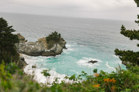 A rocky coastline with turquoise waves crashing against the shore, trees and greenery on the cliffs, and an overcast sky above. by Justin Salem Meyer
