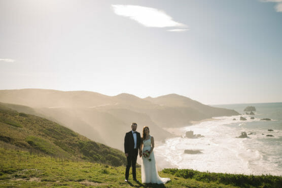 A bride and groom stand together on a grassy hill overlooking a scenic coastline, with waves crashing against rocky cliffs under a bright, cloudy sky. by Justin Salem Meyer