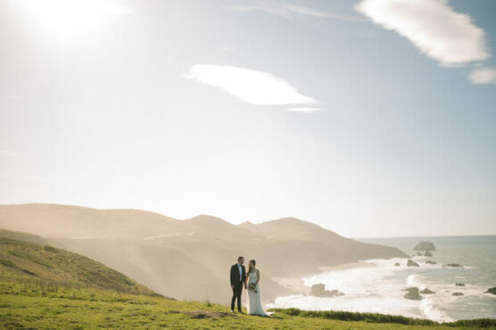 A bride and groom stand together on a grassy cliff overlooking the ocean, with rolling hills and rocky coastline in the background under a bright, partly cloudy sky. by Justin Salem Meyer
