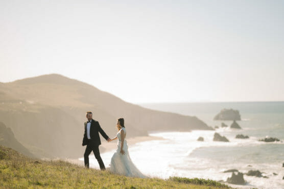 A bride and groom walk hand in hand along a grassy cliff overlooking the ocean, with waves crashing against rocky shores and hazy hills in the background under a clear sky. by Justin Salem Meyer
