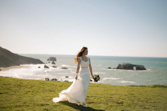 A bride in a flowing white dress holds a bouquet and stands on grassy cliffs overlooking the ocean, with waves and rocky outcrops in the background under a clear blue sky. by Justin Salem Meyer