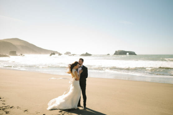 A bride and groom stand on a sandy beach near the ocean, embracing and smiling. The bride’s dress flows in the breeze, and waves and rocky outcrops are visible in the background under a clear sky. by Justin Salem Meyer