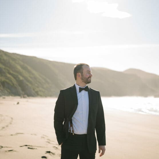 A man in a black suit and bowtie stands on a sandy beach with hills in the background, looking to the side and smiling under bright sunlight. by Justin Salem Meyer