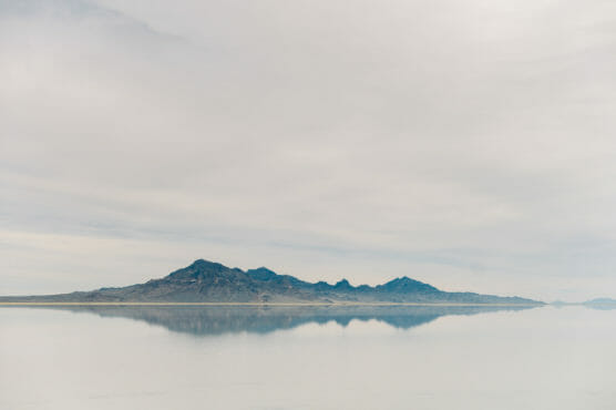 Distant mountains reflected in a calm, flat body of water under a cloudy sky, creating a serene and minimalist landscape. by Justin Salem Meyer