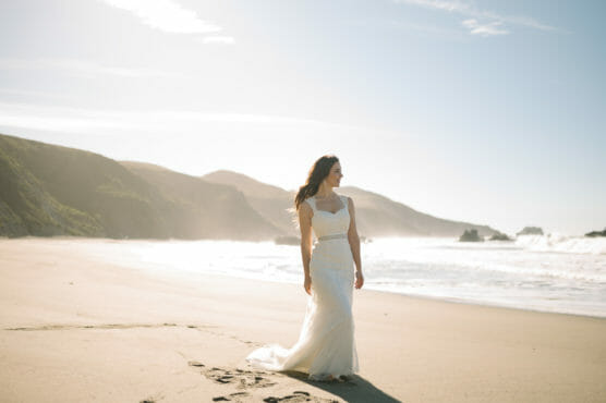 A woman in a white wedding dress stands on a sandy beach near the ocean, with waves and distant hills in the background under a bright, clear sky. by Justin Salem Meyer