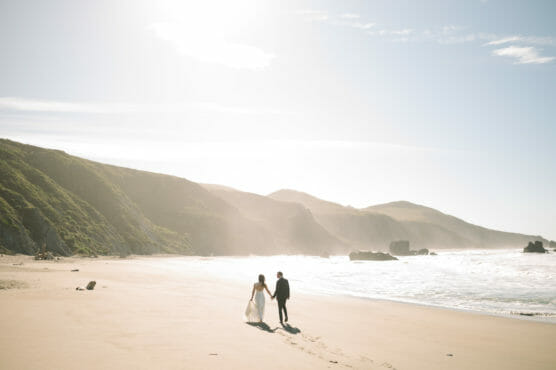 A bride and groom walk hand-in-hand along a sandy beach with waves crashing nearby and sunlit hills in the background. by Justin Salem Meyer