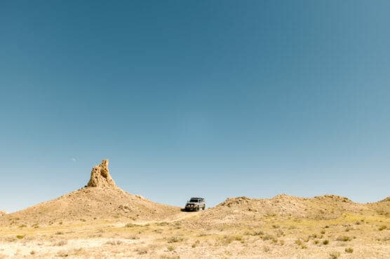 A jeep is parked on a dry, rocky hill under a clear blue sky, with a prominent rock formation rising on the left side of the landscape. Sparse vegetation covers the ground. by Justin Salem Meyer