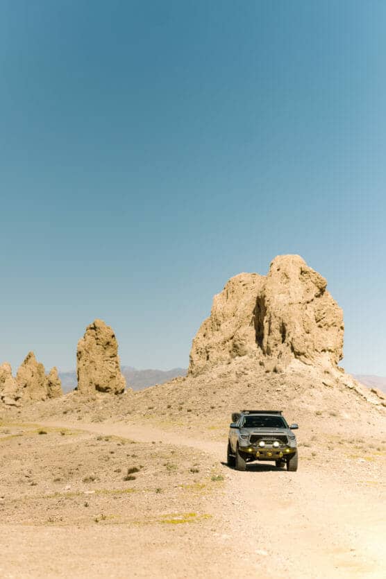 A black SUV drives on a dusty dirt road through a desert landscape with large, rugged rock formations under a clear blue sky. by Justin Salem Meyer