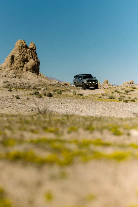 A black off-road vehicle with roof gear is parked on rocky, sandy terrain near a tall rock formation under a clear blue sky. Sparse patches of green and yellow vegetation dot the foreground. by Justin Salem Meyer