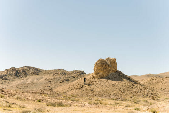 A person stands near a large rock formation in a desert landscape under a clear blue sky, surrounded by dry, hilly terrain with sparse vegetation. by Justin Salem Meyer