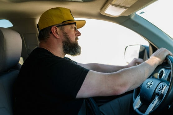 A man with a beard and glasses, wearing a yellow cap and black shirt, drives a Toyota car. He is focused on the road, holding the steering wheel with his right hand and looking out the front windshield. by Justin Salem Meyer